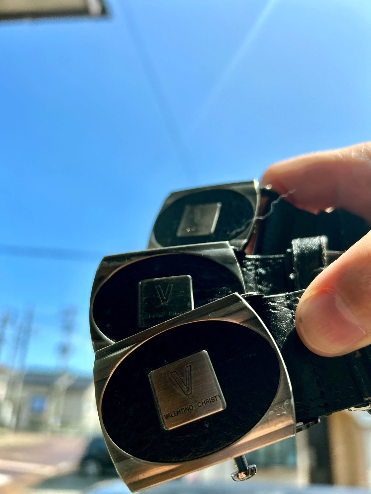 VALENTINO CHRISTY のバックルが付いた同じベルト 3 本を、青空にかざす手
